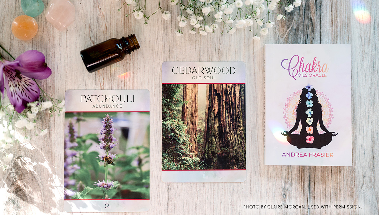 Cards from the Chakra Oils Oracle Deck displayed on a light wooden surface alongside gemstones, flowers, and an essential oil bottle. The Patchouli and Cedarwood cards sit beside the deck’s guidebook featuring a silhouette with chakra flowers.